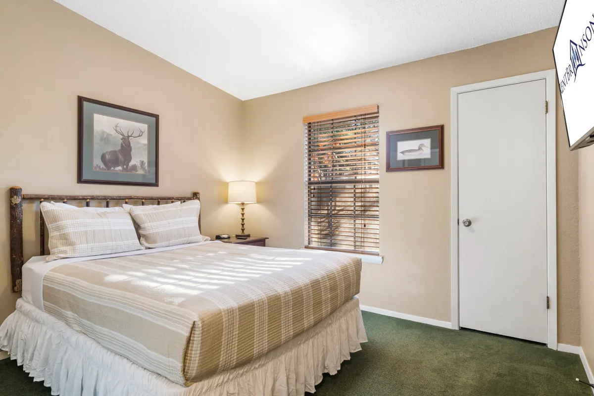This image showcases a tranquil bedroom with a neatly made bed, a bedside lamp, framed artwork, and a window letting in natural light. The bedding looks plush and inviting, and the overall decor is serene and restful.
