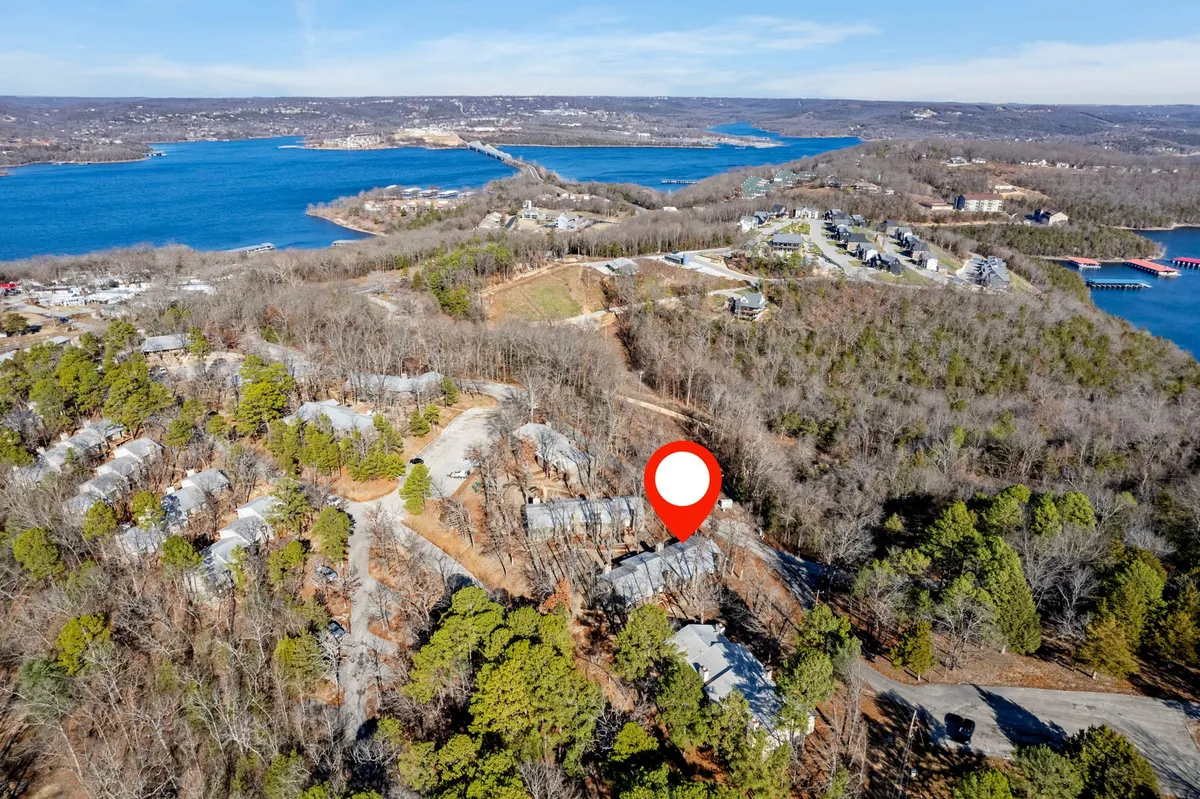 A tranquil exterior view featuring the lake surrounded by lush forest and residential structures nestled among the trees. The scene suggests accessibility to natural environments, walking areas, and the peaceful outdoors.