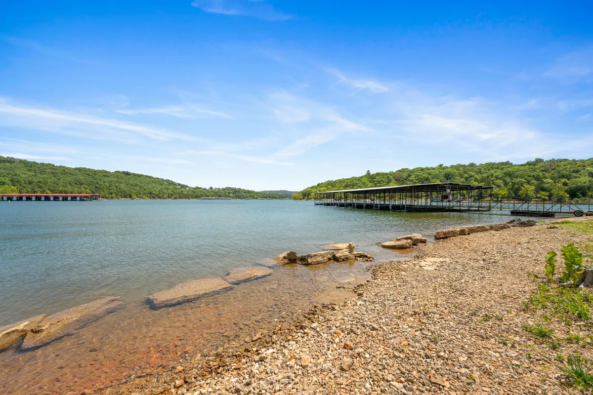 A tranquil lake scene with calm water, a boathouse, and a rocky shoreline, bordered by distant green hills. The area is perfect for boating, fishing, swimming, and picnicking, with a strong sense of waterfront relaxation.