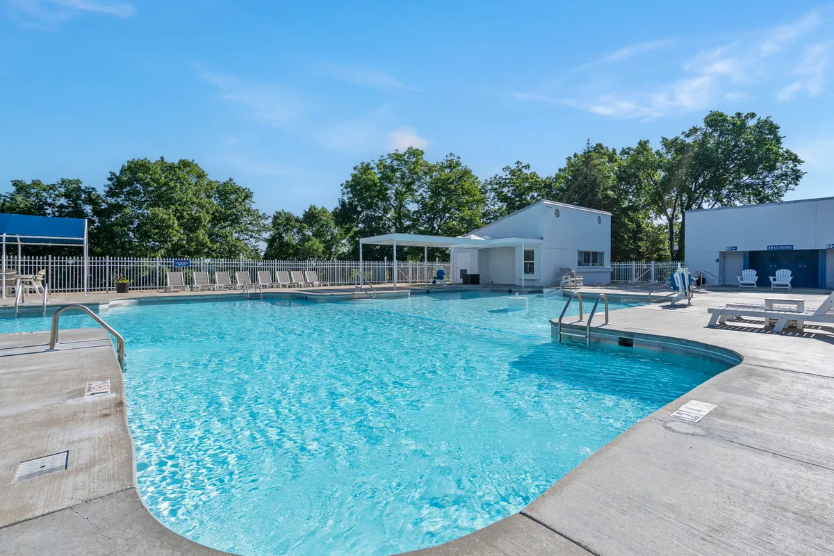 A bright, inviting swimming pool with lounge chairs and a shade structure, set against the backdrop of leafy trees. Guests are seen swimming, sunbathing, and socializing in a relaxed resort environment.