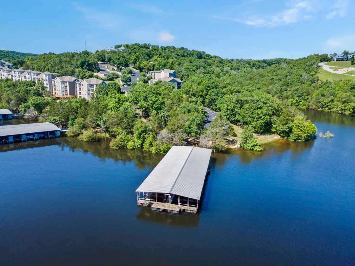 A serene lakeside view showcasing a boat dock extending over calm water, surrounded by lush greenery and residential buildings in the background. The dock is ready for mooring boats, and the inviting landscape hints at leisurely afternoons fishing, boating, or relaxing by the water.