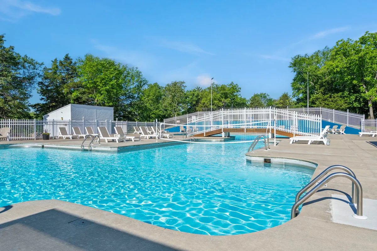 A scenic outdoor swimming pool bordered by lounge chairs and lush greenery, with a small bridge crossing over part of the pool area. The tranquil setting is ideal for relaxation or socializing on sunny days.