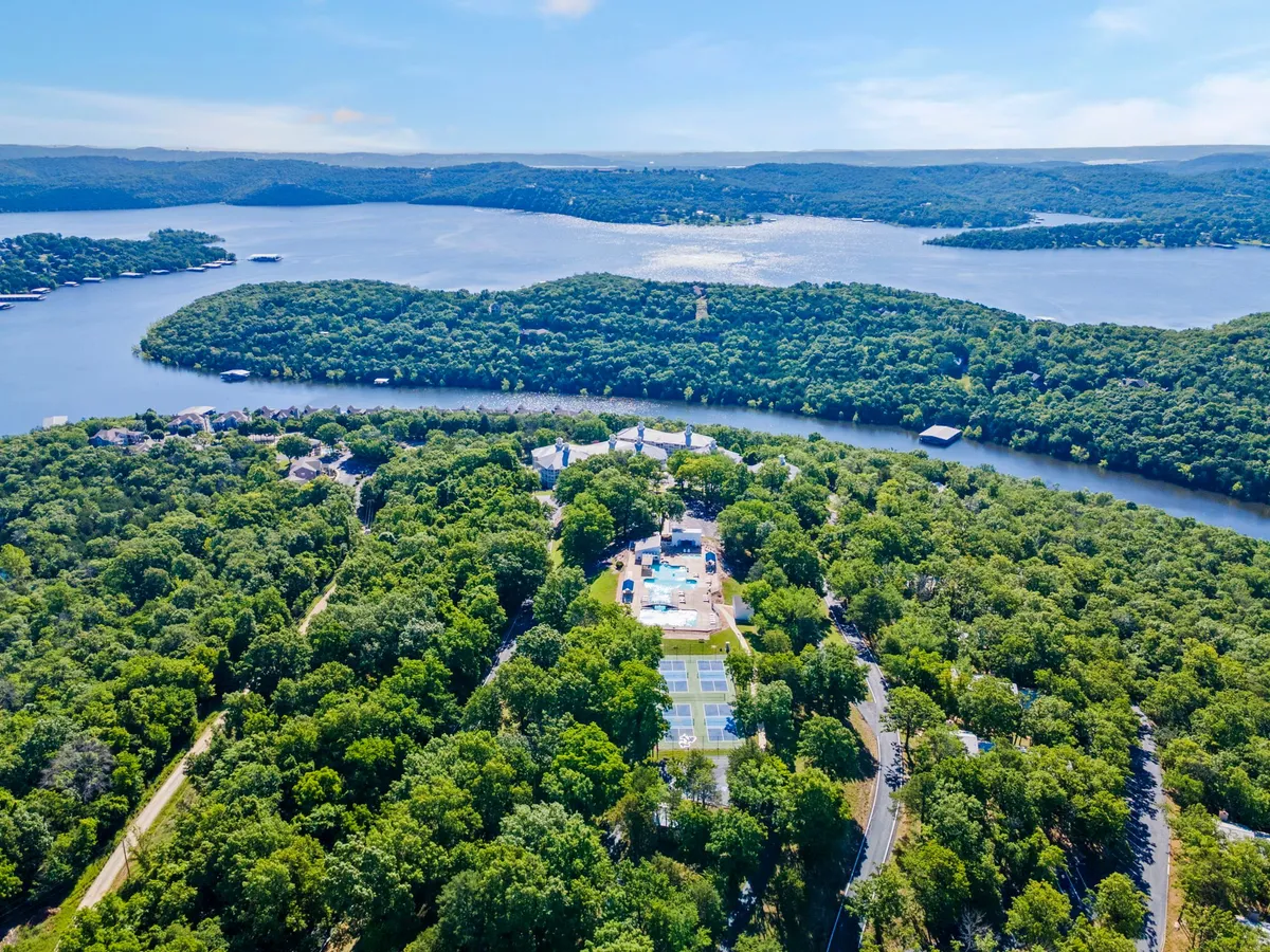This elevated view captures an expansive water body surrounded by lush green trees, with a visible swimming pool area and tennis courts in the foreground, all set against rolling hills. The image conveys both the recreational amenities and the natural setting of the resort.