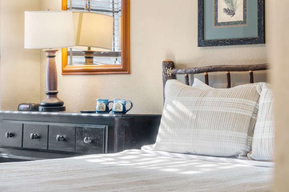 A second view of a bedroom featuring a comfortable bed with striped pillows, a nightstand with coffee mugs, a lamp, and tasteful wall art. The natural light and thoughtful details create an inviting, restful mood.